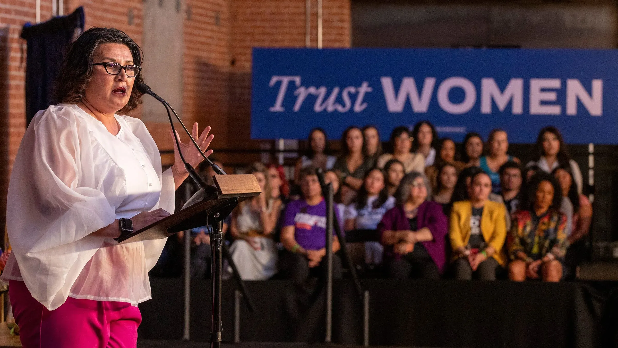 امرأة تتحدث من على المنصة في حدث لدعم حقوق المرأة، مع لافتة كبيرة خلفها تحمل عبارة "ثق بالنساء"، وحضور كبير من النساء في الجمهور.
