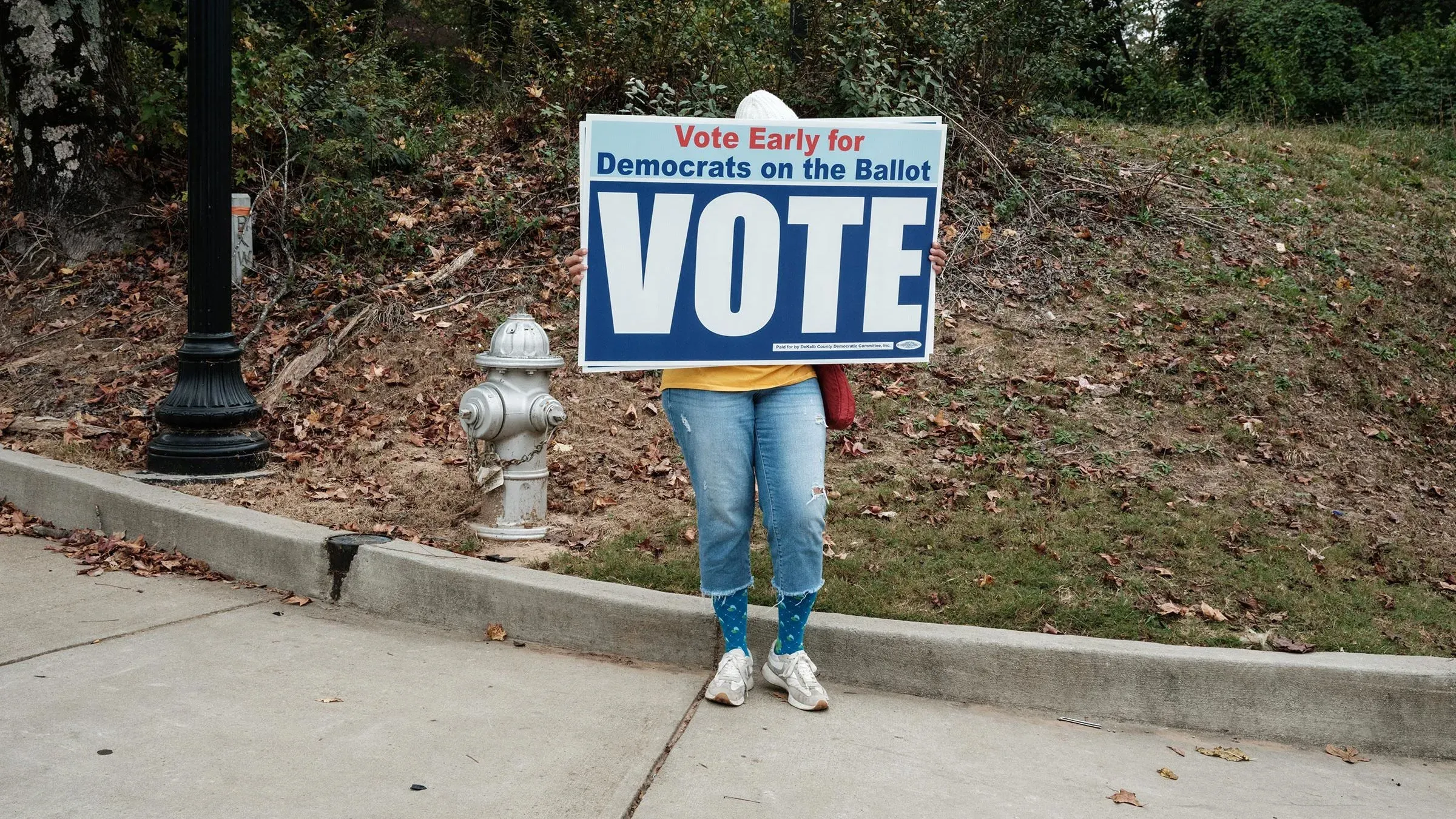 امرأة تحمل لافتة كبيرة تدعو للتصويت المبكر لصالح الديمقراطيين، وسط طبيعة خريفية. تعكس الصورة أهمية المشاركة السياسية في الانتخابات المقبلة.