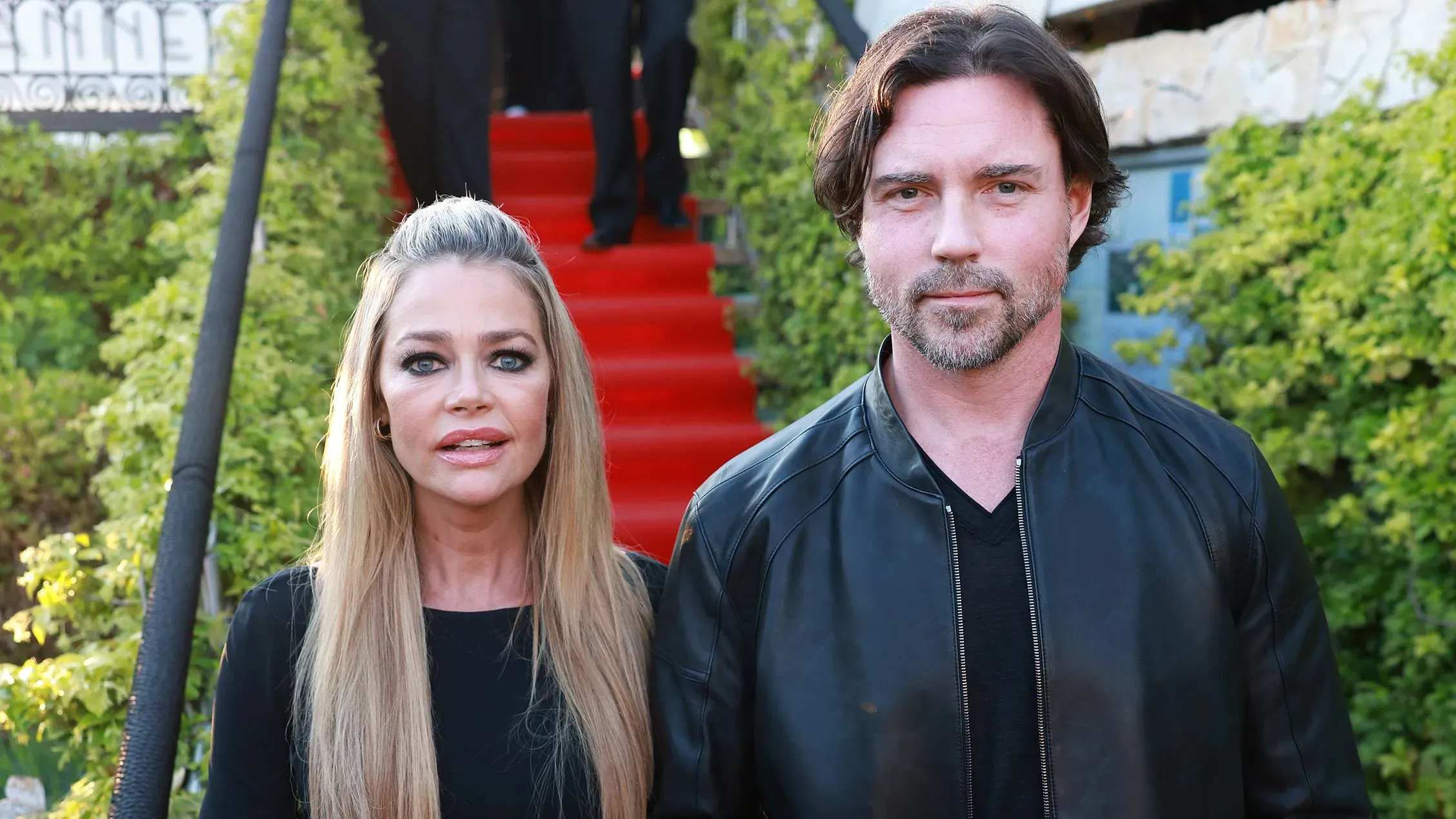 دينيس ريتشاردز وآرون فيبرز يقفان معًا في حدث عام، حيث تظهر ريتشاردز بوضوح مع علامات على وجهها تشير إلى إصابة.