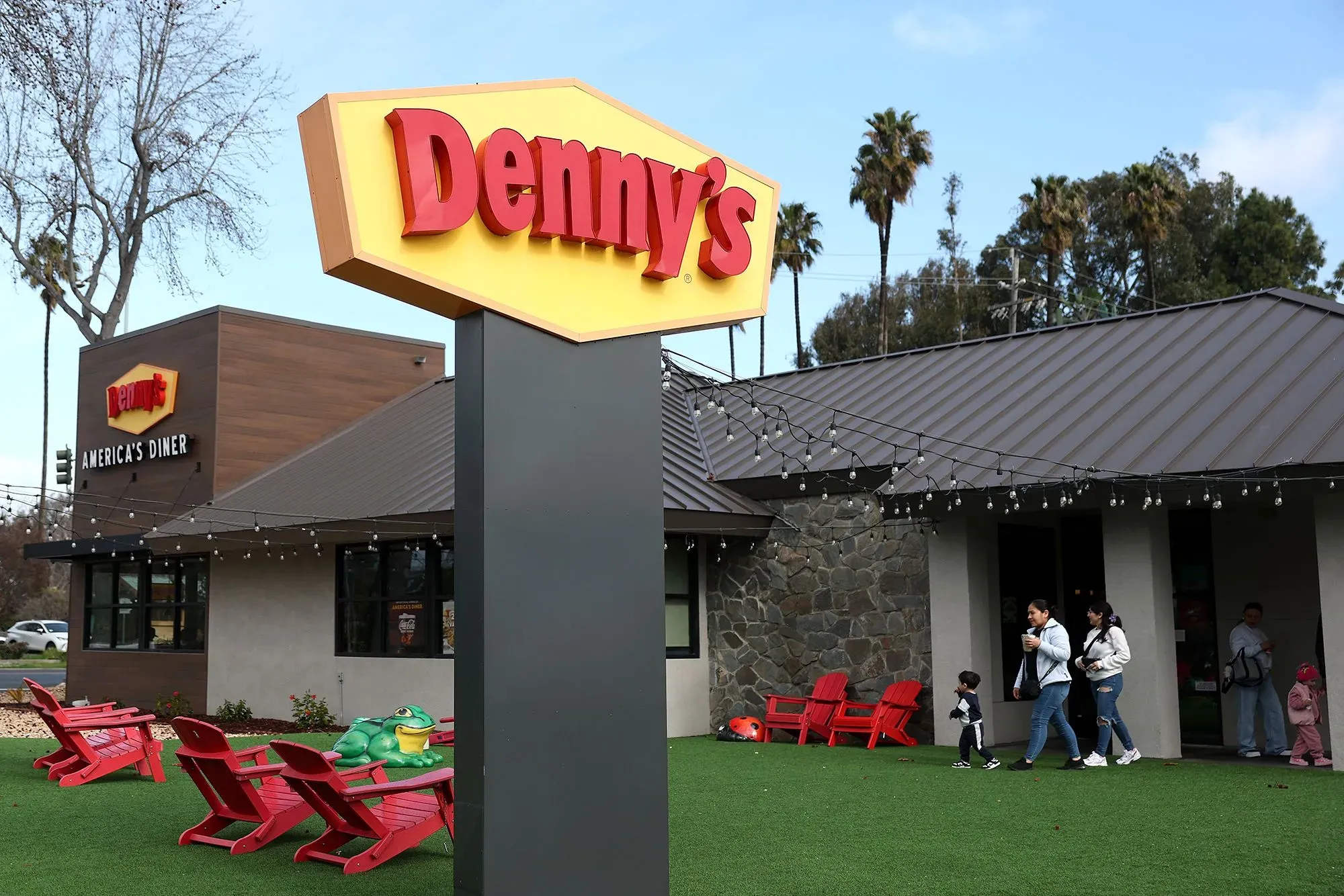 لافتة مطعم دينيز تظهر اسم المطعم "Denny's" مع عائلات تتجول في الخارج، تعكس التحديات التي تواجهها السلسلة في السوق.