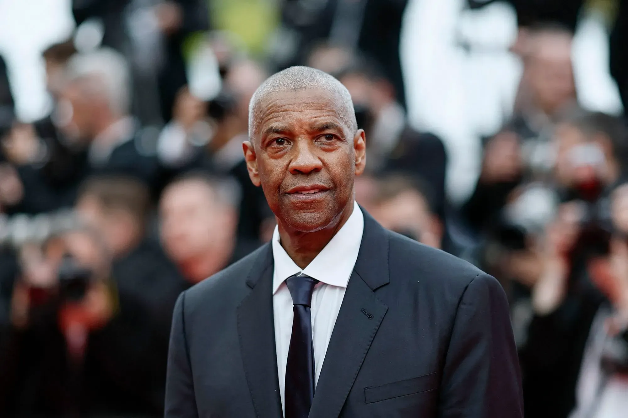 دينزل واشنطن في عرض رسمي، يرتدي بدلة أنيقة، وسط حشد من المصورين، مع التركيز على تعبيره الجاد أثناء مناقشة صناعة الأفلام.