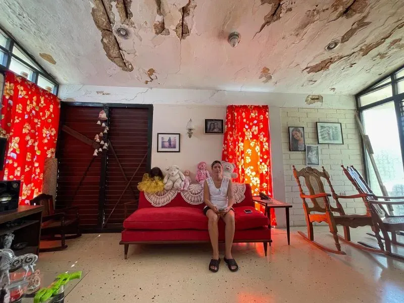 امرأة تجلس على أريكة حمراء في غرفة معيشة متهالكة، محاطة بصور ودمى، تعكس ظروف الحياة الصعبة في كوبا بعد الترحيل.