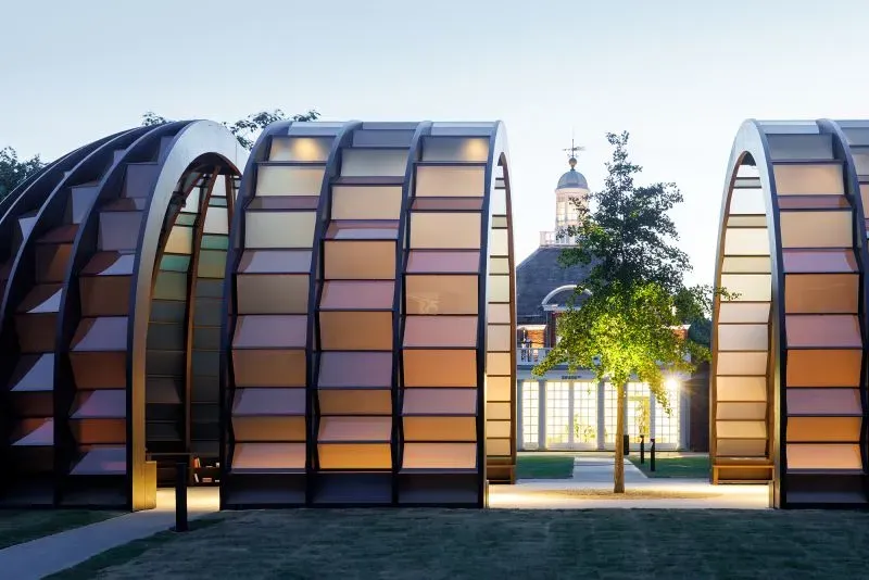 تصميم جناح سربنتين لعام 2023 يظهر هيكلًا خشبيًا منحنيًا مع ألواح شفافة، محاطًا بشجرة جينكو، يعكس تأثير الضوء في المساحات المعمارية.