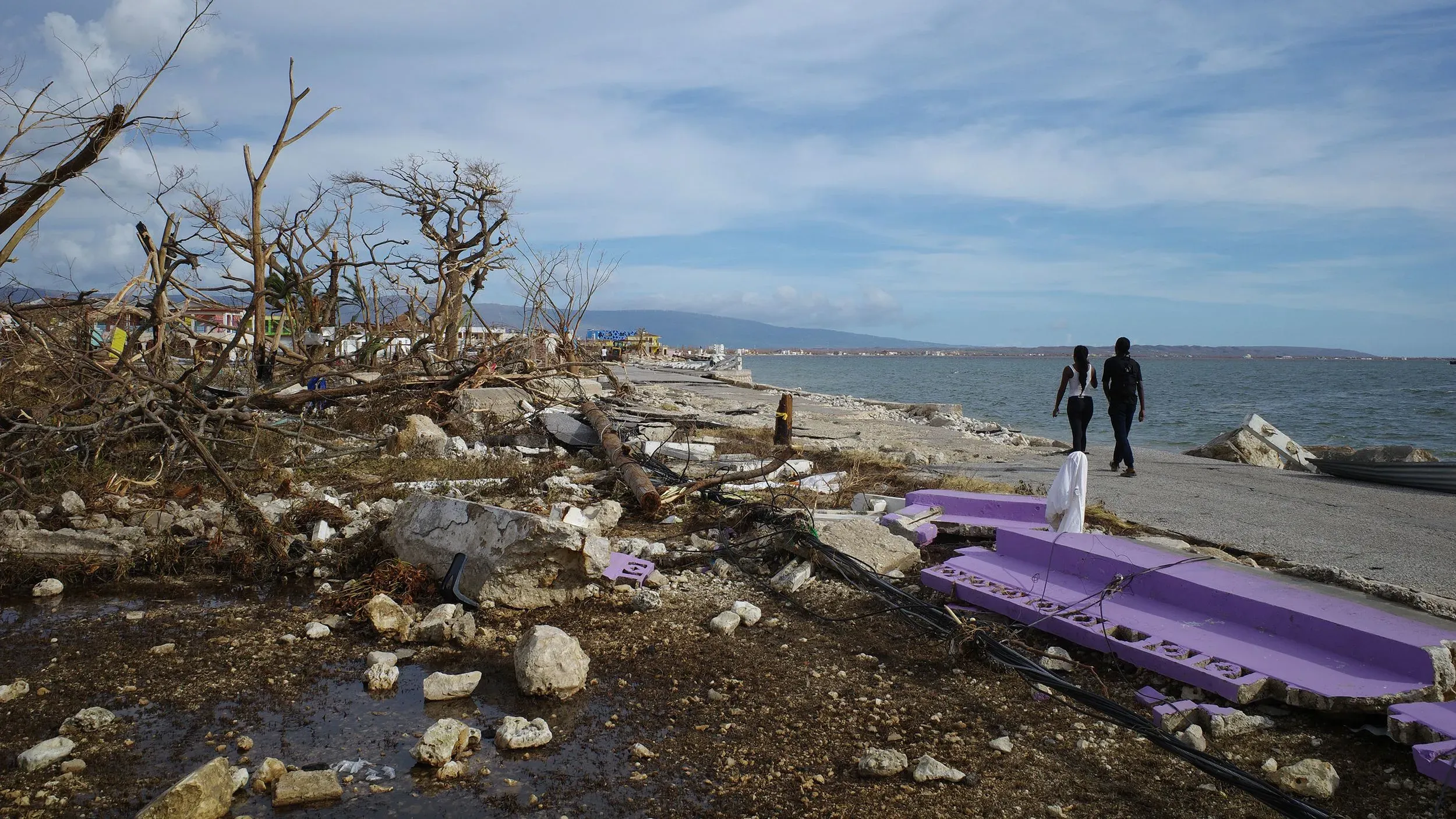 تظهر الصورة مشهدًا مدمّرًا في جامايكا بعد إعصار ميليسا، مع بقايا المنازل والأشجار المكسورة، بينما يسير شخصان على الواجهة البحرية.