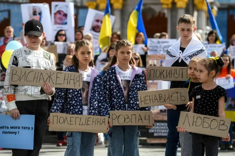 أطفال أوكرانيون يحتجون في الشارع، يحملون لافتات تعبر عن مشاعر الفقد والحنين، وسط صور لمحتجزين في الخلفية.