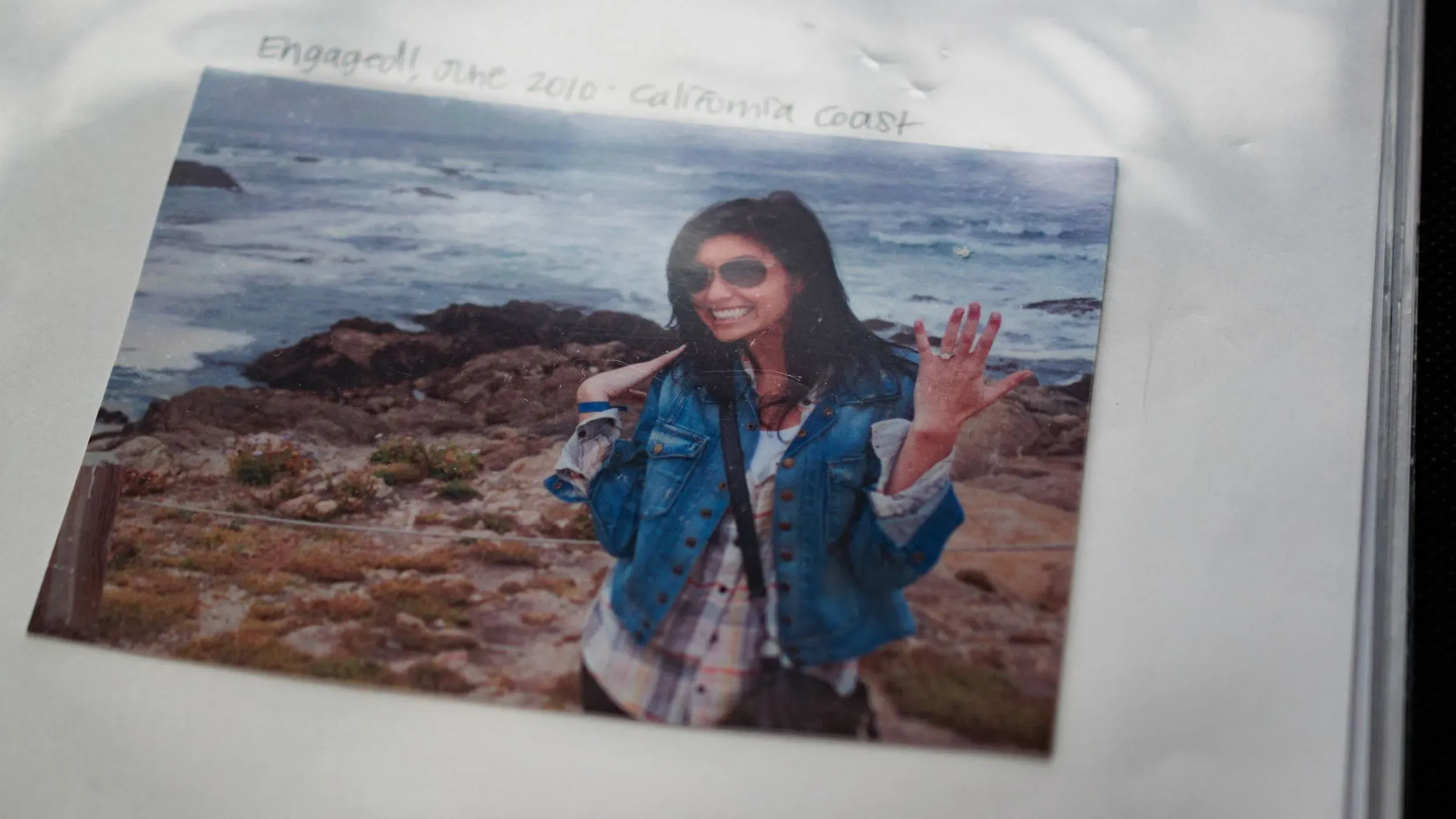 صورة لإيلين غرينبرغ، معلمة مدرسة، مبتسمة وترتدي سترة زرقاء، تشير بإصبعها إلى خاتم الخطوبة، بينما تظهر خلفها شواطئ كاليفورنيا.