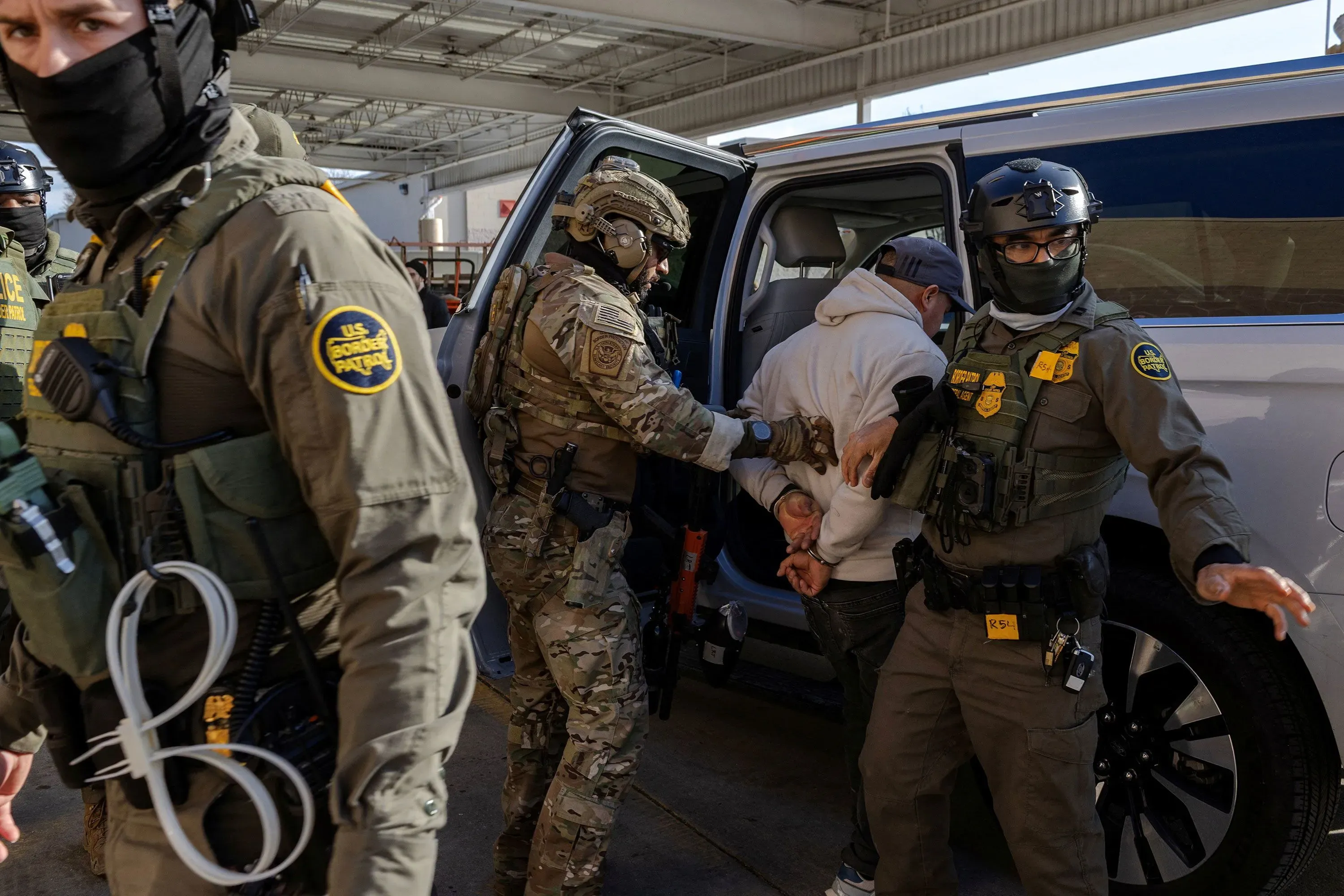 اعتقال مهاجر من قبل ضباط الهجرة في محطة وقود، مع وجود عناصر أمنية مسلحين في الخلفية، يعكس التوترات حول سياسات الهجرة.