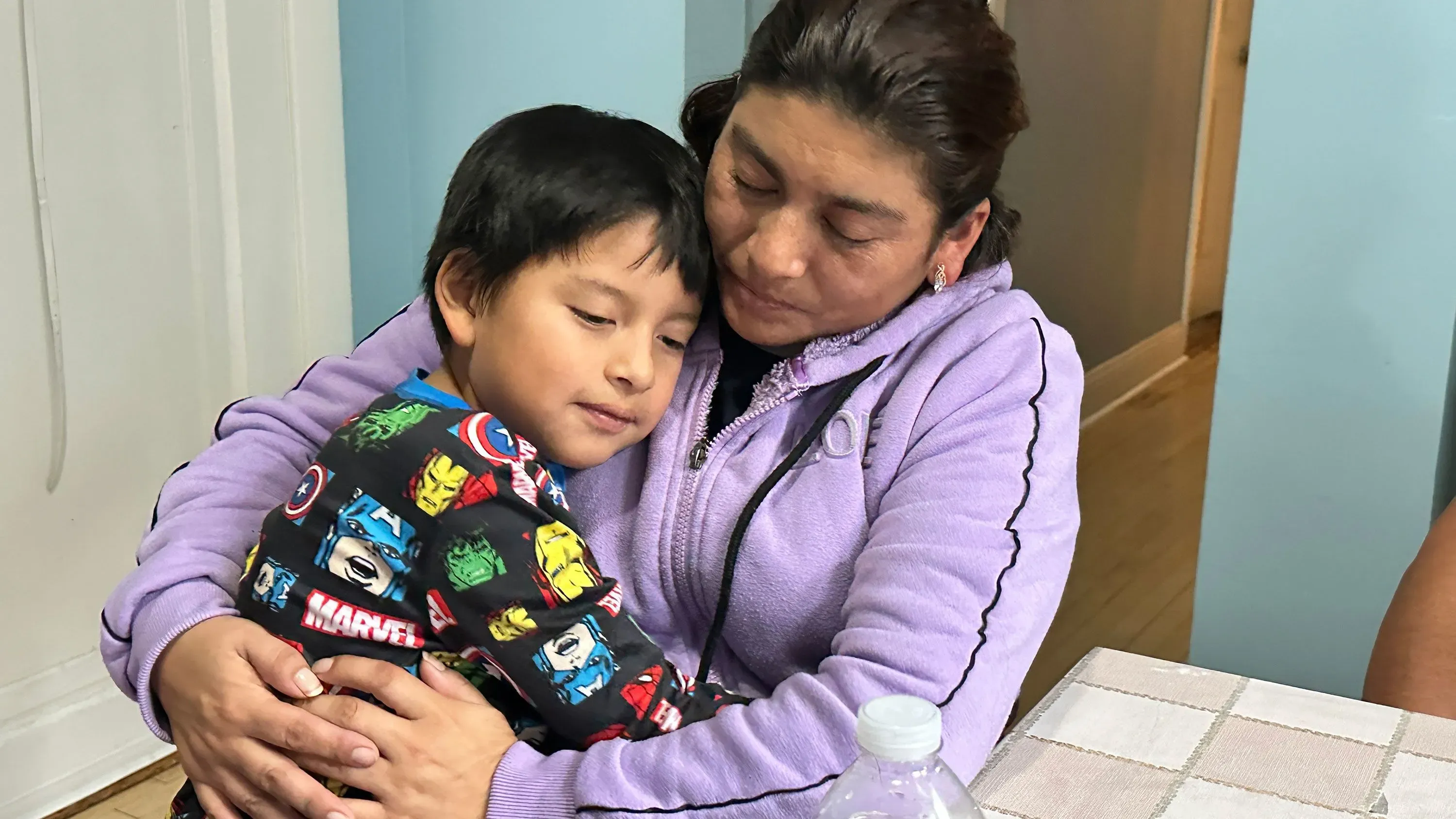 امرأة تحتضن طفلًا صغيرًا في غرفة منزلية، تعكس مشاعر الحنان والقلق وسط قضايا الهجرة.