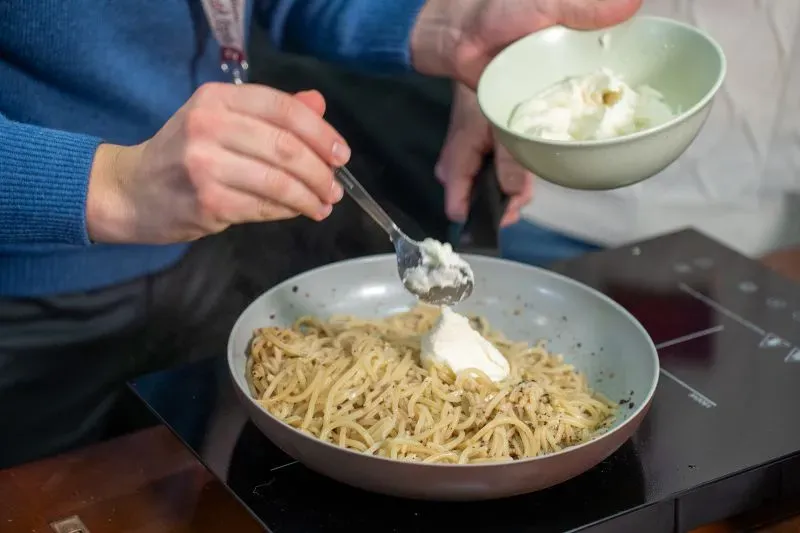 شخص يضيف جبنة كريمة إلى طبق من المعكرونة أثناء تحضير وصفة كاسيو إي بيبي، مع التركيز على تقنيات الطبخ الدقيقة.