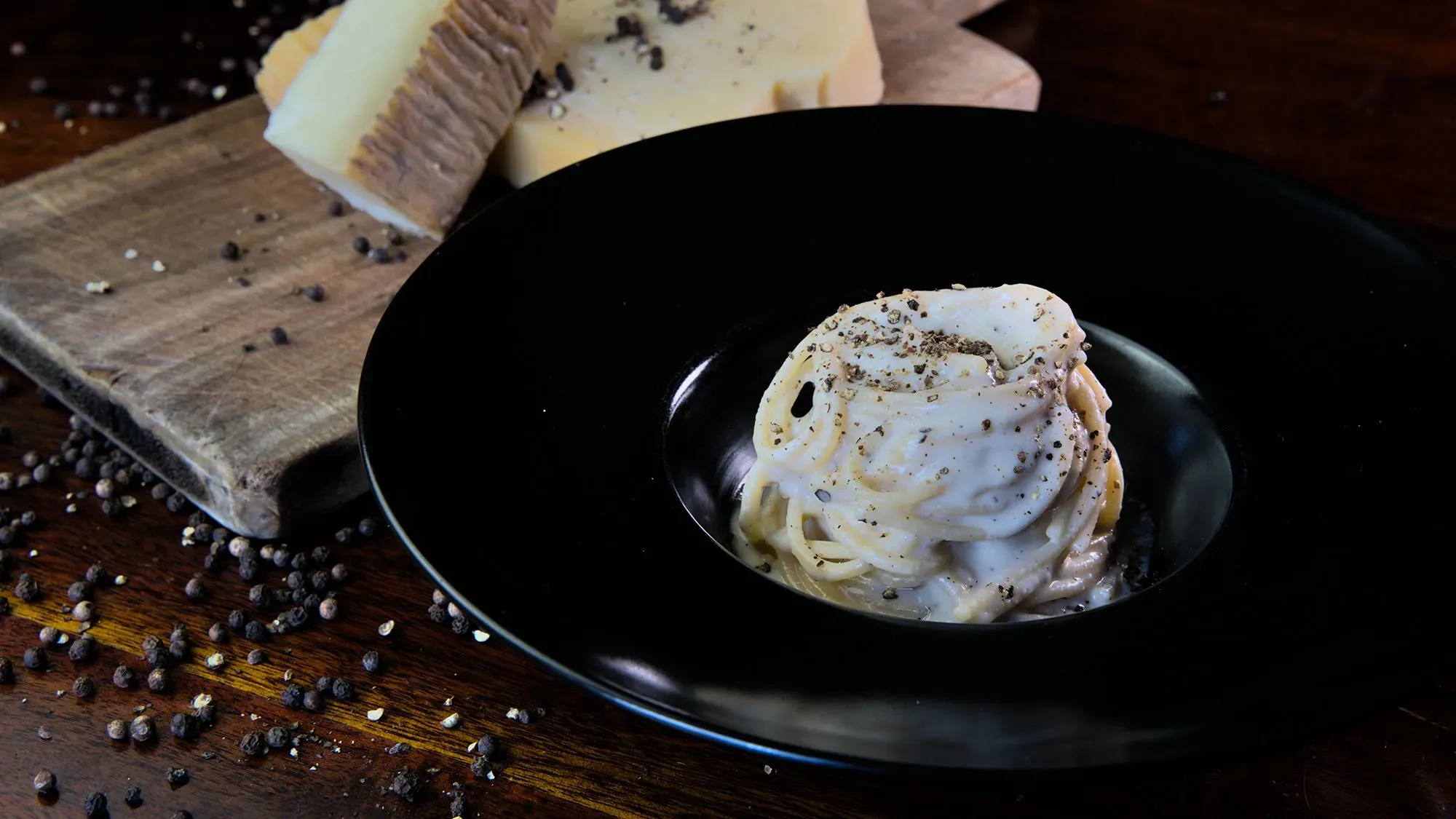 طبق المعكرونة كاسيو إي بيبي مزين بالفلفل الأسود، مع قطع من جبن بيكورينو رومانو، يقدم في وعاء أسود مع حبيبات فلفل حوله.