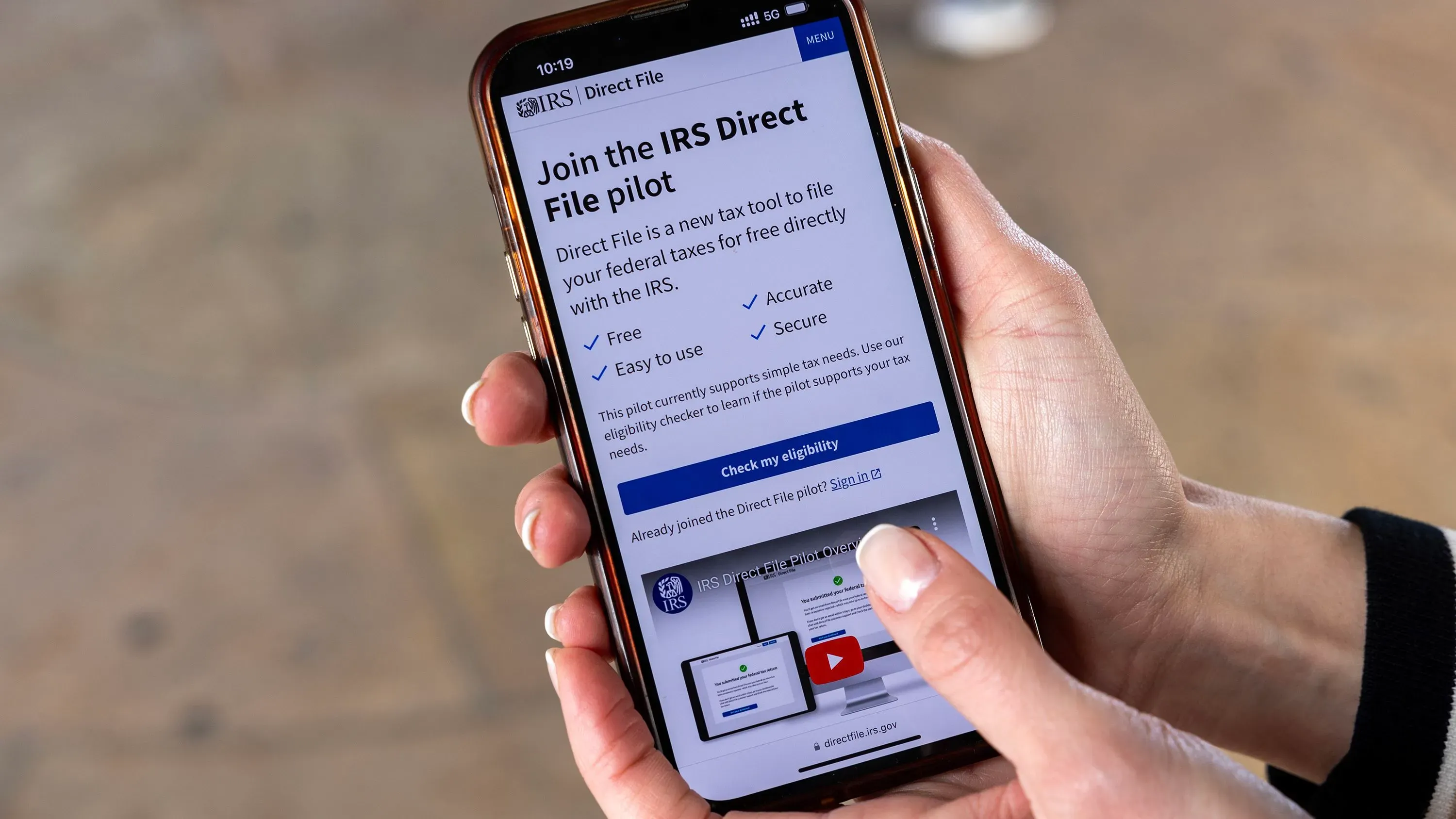 تظهر الصورة شخصًا يحمل هاتفًا ذكيًا يعرض صفحة برنامج "IRS Direct File" لتقديم الإقرارات الضريبية مجانًا وسهولة الاستخدام.