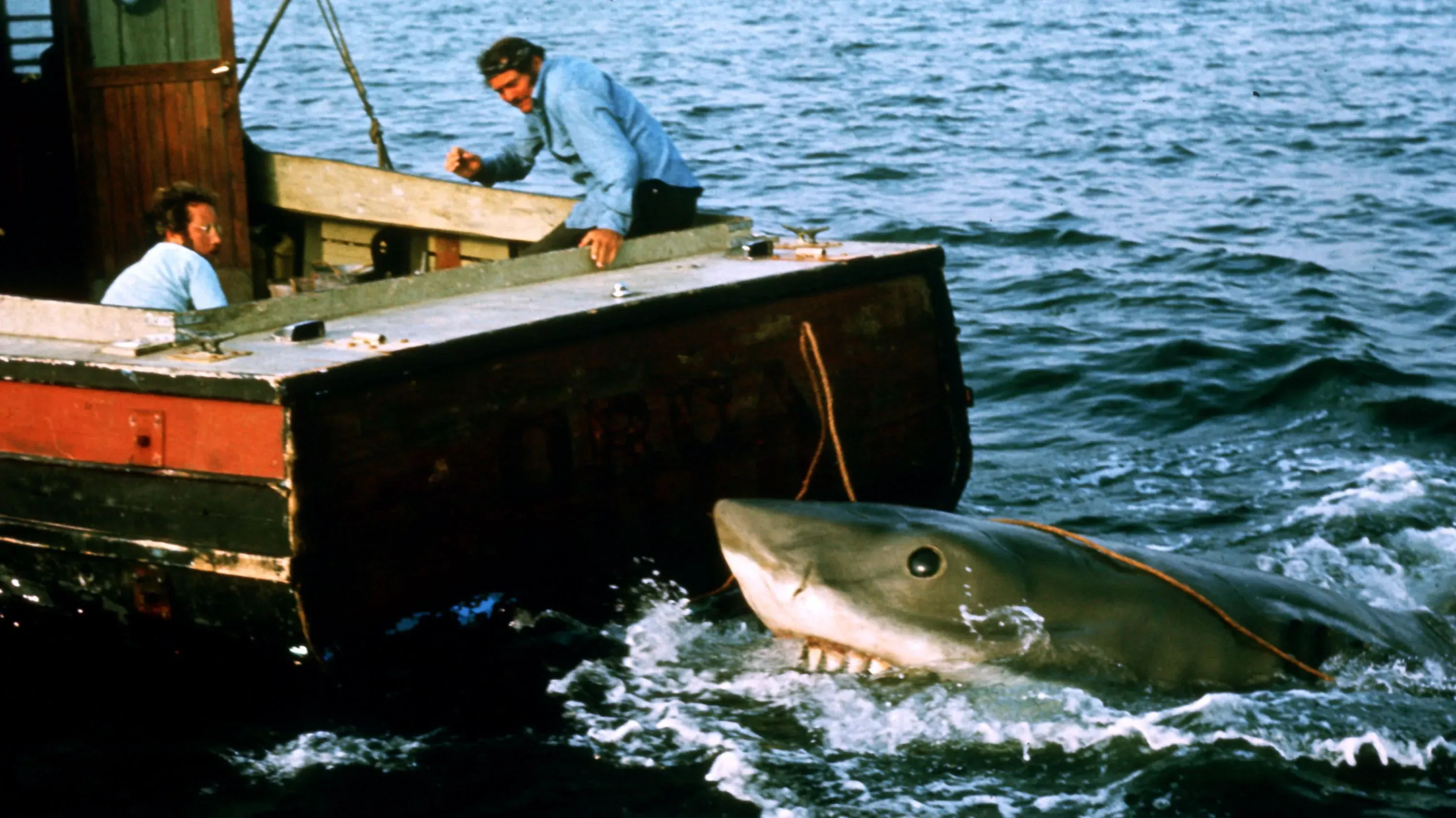 صورة تظهر سمكة قرش بيضاء كبيرة تقترب من قارب، حيث يتواجد رجلان على متنه، مما يعكس مشهدًا من فيلم "الفك المفترس" وتأثيره على ثقافة الخوف من أسماك القرش.