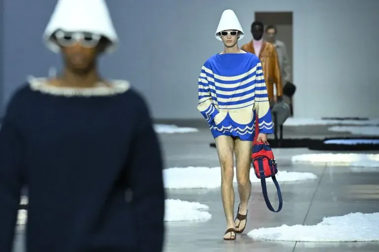 عارض أزياء يرتدي سترة زرقاء مخططة وشورت قصير، يسير على منصة عرض الأزياء في أسبوع الموضة بميلانو، مع خلفية فنية.