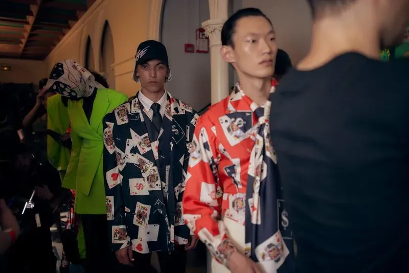 عرض أزياء رجالية في باريس، يظهر عارضون يرتدون سترات مزينة برسومات بطاقات لعب وألوان زاهية، مع أجواء حيوية خلفهم.