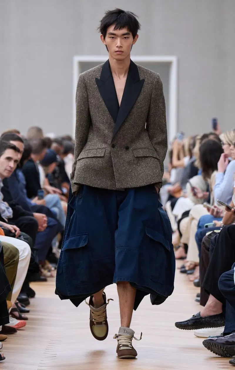 عارض شاب أزياءً مبتكرة في عرض أزياء باريس، مرتديًا سترة مزدوجة الصدر وسروالًا فضفاضًا، أمام جمهور من النخبة.