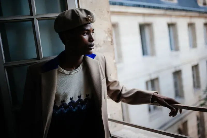 رجل يرتدي سترة أنيقة وقبعة، يقف بجانب نافذة في موقع عرض أزياء باريس، مع خلفية معمارية مميزة.