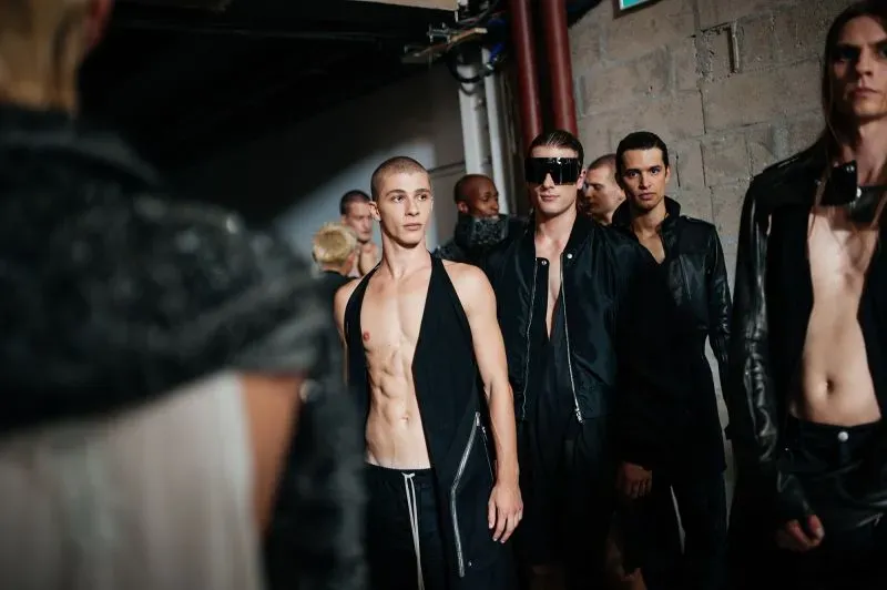 عرض لعارضين يرتدون أزياء عصرية في كواليس أسبوع الموضة في باريس، مع التركيز على التصاميم الجريئة والألوان الداكنة.