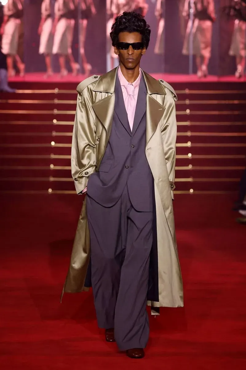 عارض يرتدي معطفًا فضيًا وسروالًا واسعًا، يسير على منصة عرض الأزياء في باريس، وسط أجواء حماسية وأضواء ساطعة.