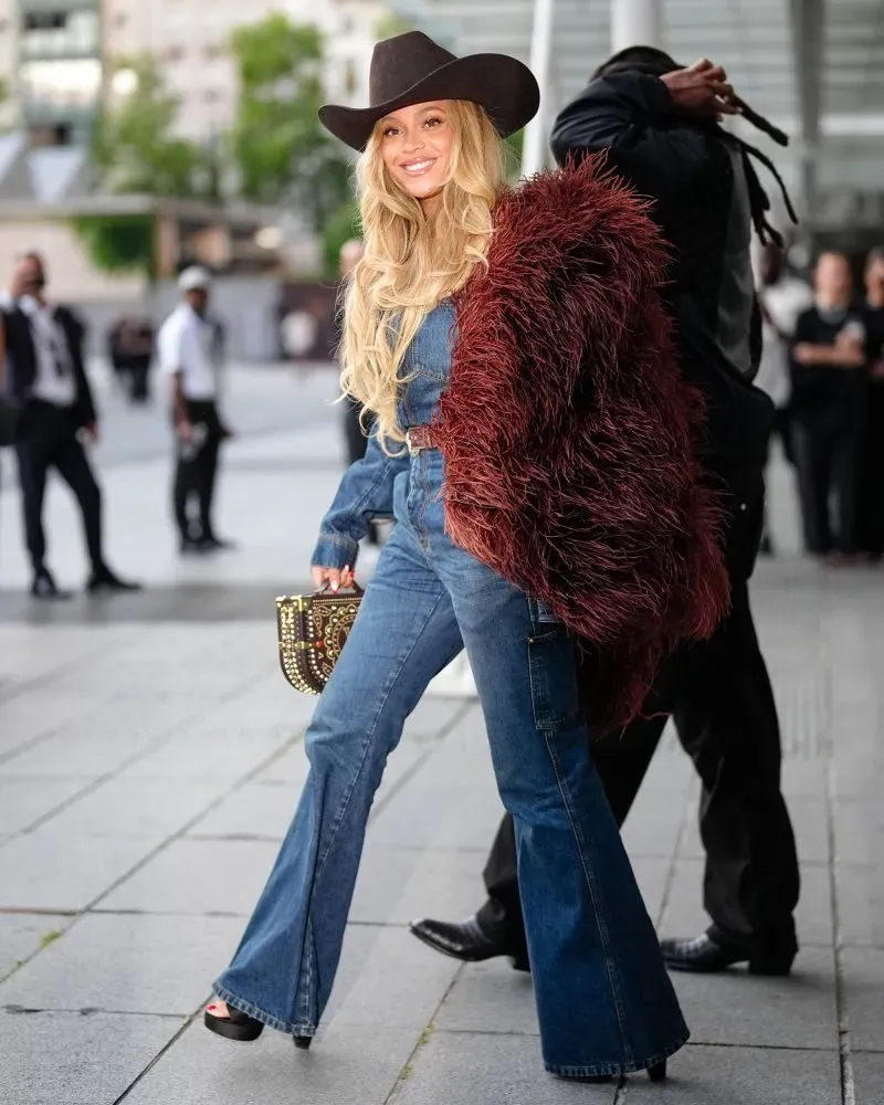 امرأة ترتدي ملابس جينز واسعة مع سترة فروية وقبعة، تحمل حقيبة يد مزخرفة، مبتسمة أمام مجموعة من الحضور في عرض أزياء باريسي.