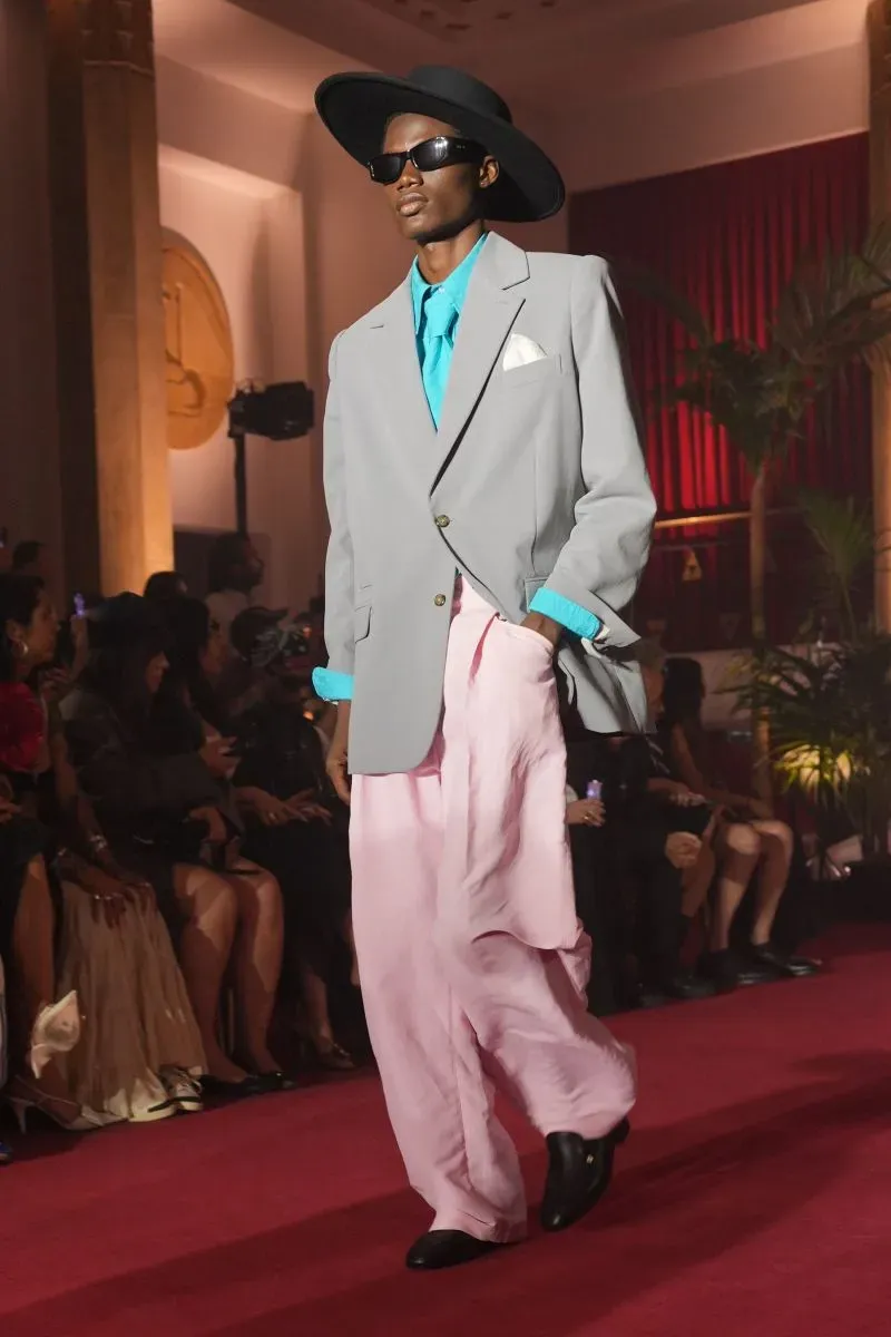 عارض يرتدي بدلة رمادية مع قميص أزرق وسروال وردي، يسير على ممشى عرض الأزياء وسط جمهور في فندق إنفاليد بباريس.