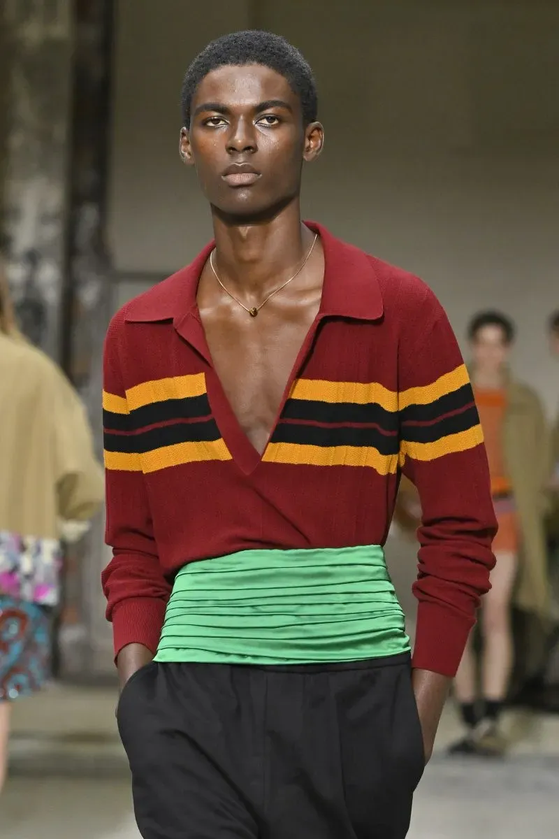 عارض أزياء شاب يرتدي سترة حمراء مخططة بألوان زاهية وحزام أخضر، يظهر في عرض أزياء باريسي يركز على الأزياء القابلة للتكيف.
