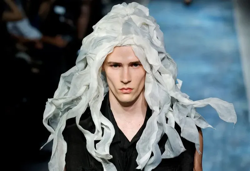 عارض أزياء شاب يرتدي تصميماً مبتكراً يتضمن غطاء رأس أبيض يشبه الشعر، يعكس أسلوباً جريئاً وأنيقاً في عرض الأزياء.
