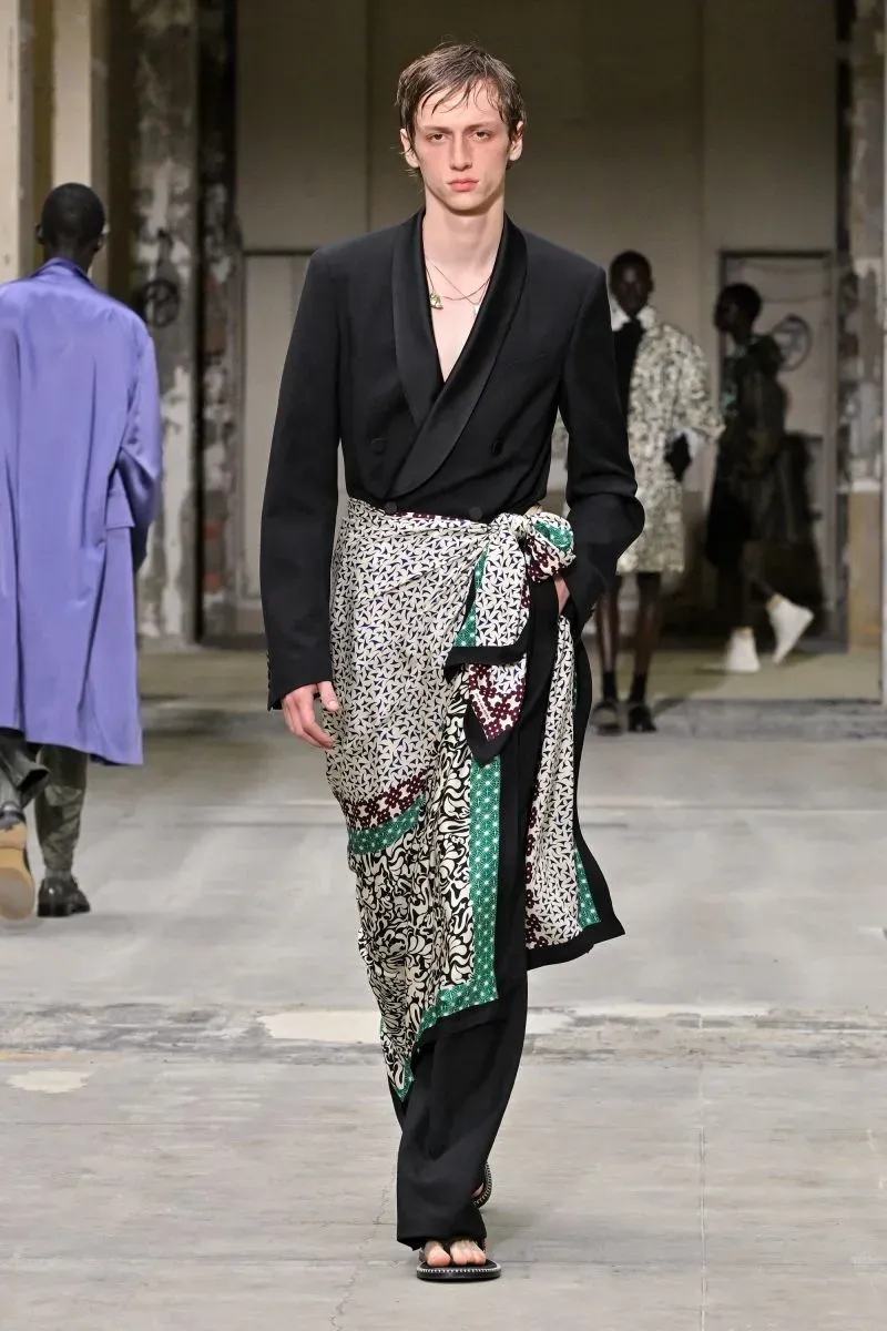 عارض شاب يرتدي بدلة سوداء مع غطاء ملون مزخرف خلال عرض أزياء في باريس، مع خلفية تعكس أجواء الحدث.