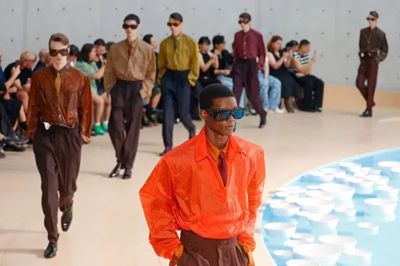 عرض أزياء رجالية في باريس، مع عارضين يرتدون ملابس ملونة عصرية ونظارات شمسية، بينما يتابع الجمهور من الخلف.