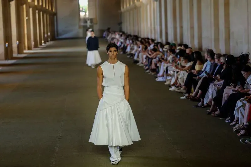 عرض أزياء رجالية في باريس، يُظهر عارضًا يرتدي فستانًا أبيضًا بتصميم مبتكر، مع جمهور متنوع في الخلفية.