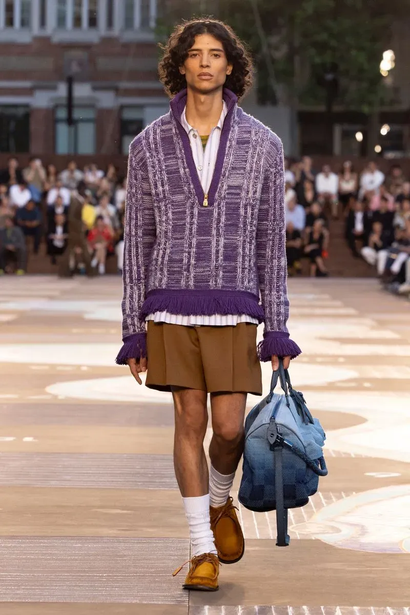 عارض شاب يرتدي سترة أرجوانية طويلة وسروال قصير بني، حاملاً حقيبة زرقاء، خلال عرض أزياء في باريس، وسط جمهور متنوع.