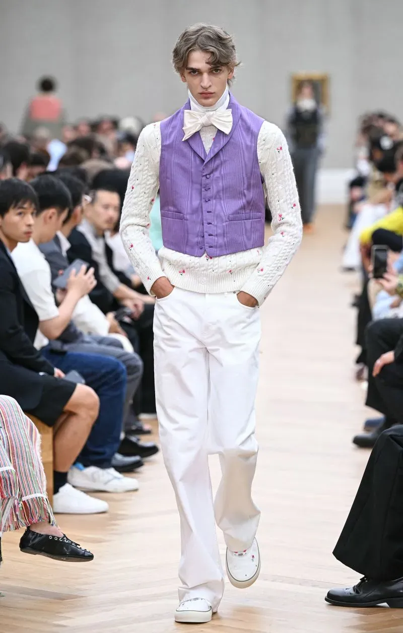 عارض أزياء شاب يرتدي سترة بنفسجية وقميصًا أبيض مزخرفًا، يسير على ممشى عرض أزياء في باريس وسط جمهور متنوع.
