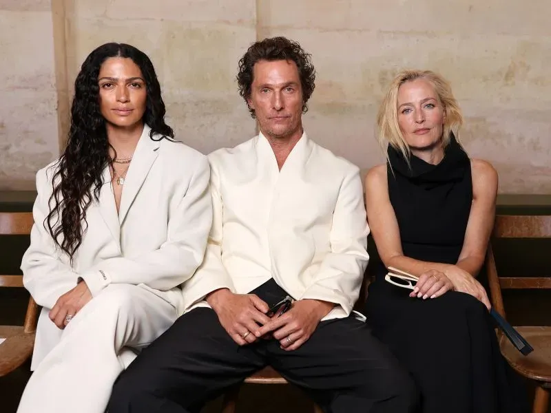 ثلاث شخصيات جالسة في عرض أزياء، يرتدون ملابس أنيقة بألوان محايدة، مع خلفية بسيطة، تعكس أجواء أسبوع الموضة في باريس.