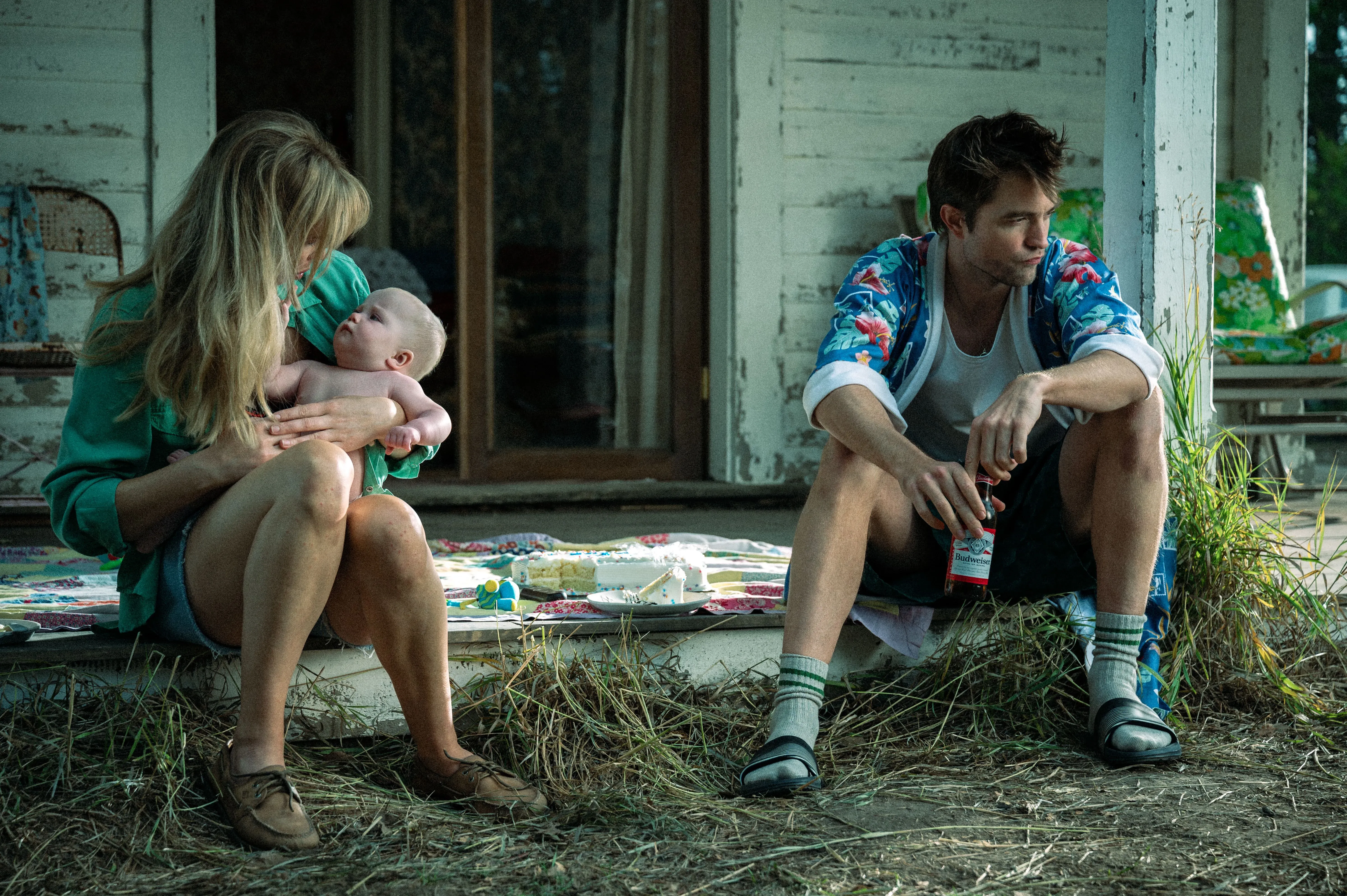 امرأة تحمل طفلاً في حضنها، بينما يجلس رجل بجانبها على عتبة منزل ريفي. تعكس الصورة التوتر في علاقتهما وسط مشهد طبيعي.