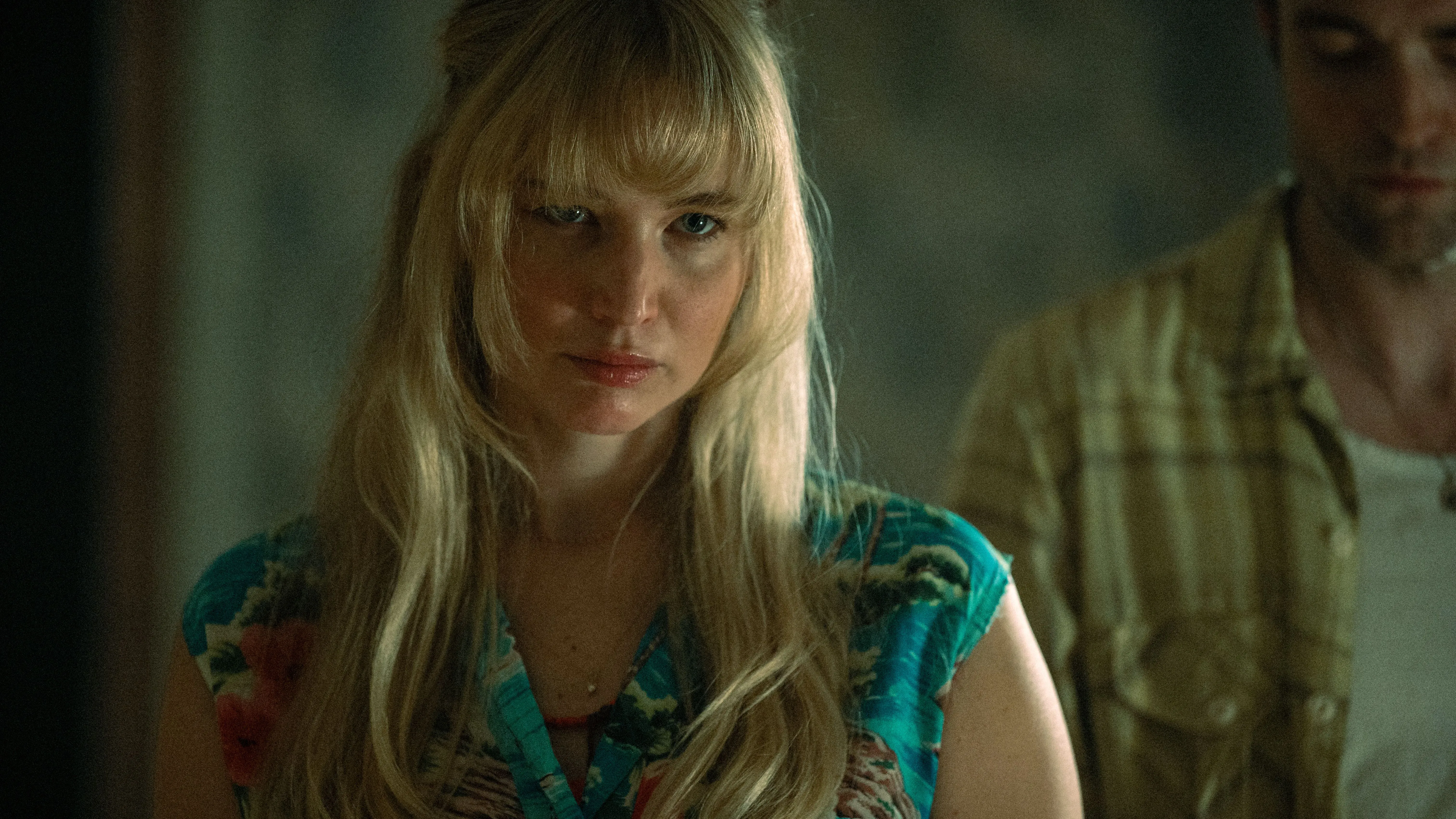 امرأة ذات شعر أشقر طويل ترتدي قميصًا مزخرفًا، تبدو عابسة بينما يقف رجل خلفها. تعكس الصورة توتر العلاقة بين الشخصيتين في فيلم "موت حبي".