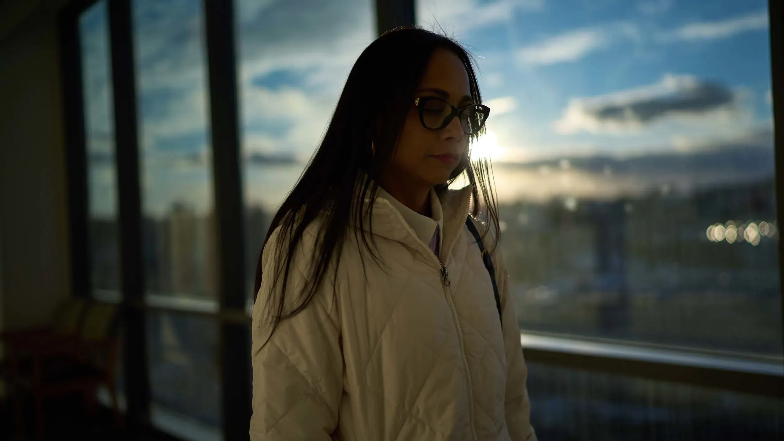 امرأة ترتدي سترة بيضاء، واقفة بجانب نافذة كبيرة، مع غروب الشمس في الخلفية، تعبر عن مشاعر القلق بشأن مرض الذئبة.