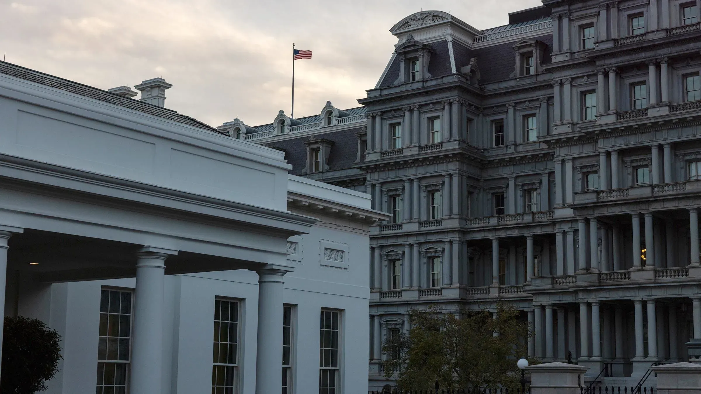 واجهة البيت الأبيض مع علم الولايات المتحدة يرفرف في الخلفية، تعكس أجواء التوتر السياسي حول تمويل الحكومة.