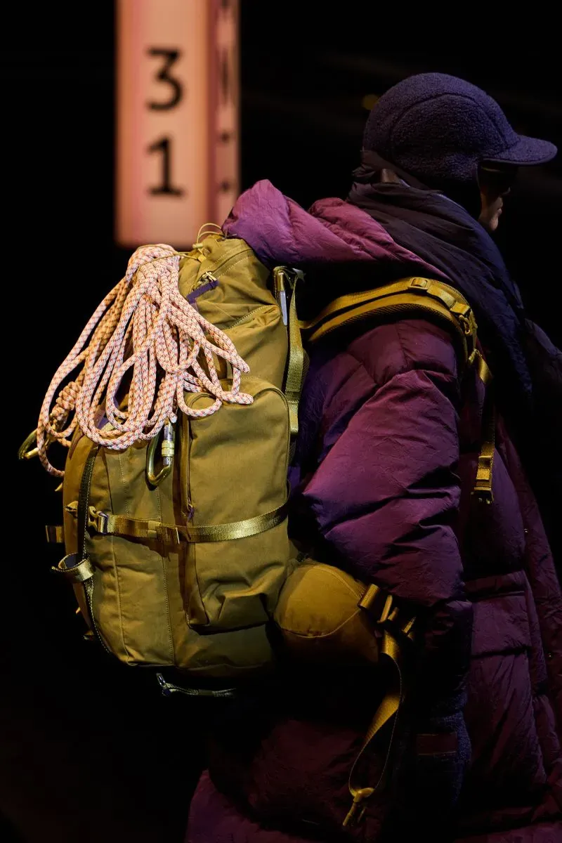 صورة تبرز جانبًا من عرض أزياء ميلانو، حيث يرتدي عارض أزياء سترة مريحة ويحمل حقيبة ظهر ذات حبال، تعكس الاتجاهات الحديثة في الملابس الخارجية.