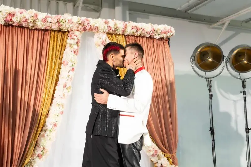 قبلة تجمع بين شخصين يرتديان ملابس ملونة وأنيقة، مع خلفية مزينة بالزهور والستائر، تعكس أجواء عروض الأزياء الحديثة.