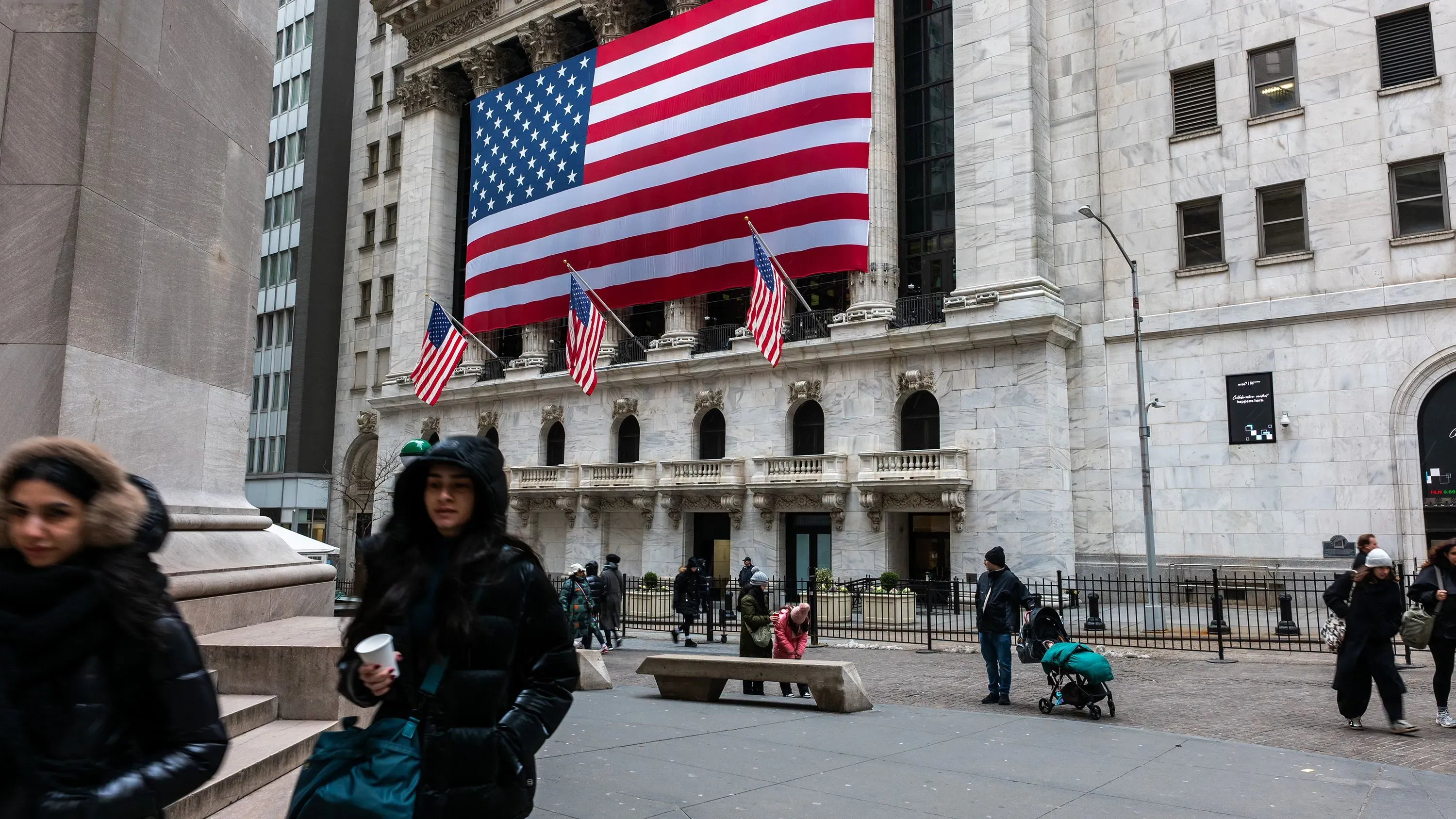 تظهر الصورة واجهة بورصة نيويورك مع علم الولايات المتحدة الكبير، بينما يتجول الناس في الشارع، مما يعكس حالة الأسواق المالية.
