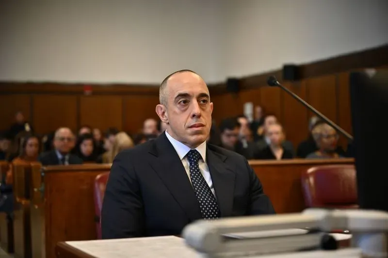 رجل يجلس في قاعة محكمة، يبدو جادًا، مع وجود جمهور خلفه. تعكس الصورة أجواء التوتر حول قضايا الفساد السياسية في نيويورك.