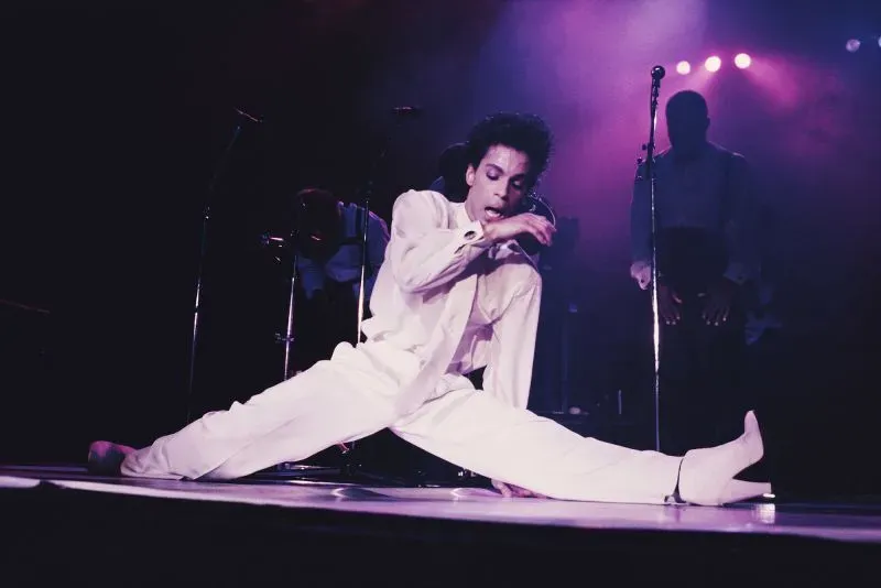 مغني يرتدي بدلة بيضاء وحذاءً بكعب عالٍ، يؤدي عرضًا موسيقيًا على المسرح مع إضاءة ملونة، مستعرضًا أسلوبه الفريد.