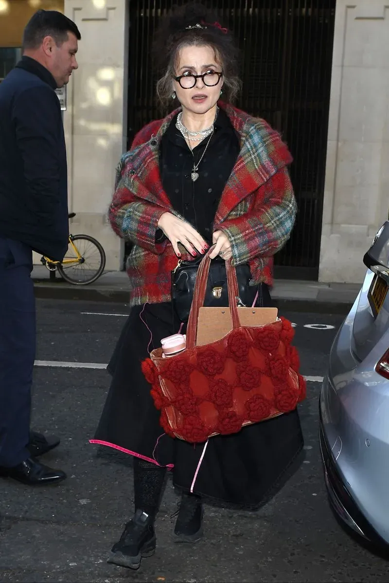 امرأة ترتدي ملابس عصرية غير متناسقة، تحمل حقيبة كبيرة، تمشي في الشارع مع رجل خلفها، تعكس أسلوب "المرأة الإنجليزية المرهقة".