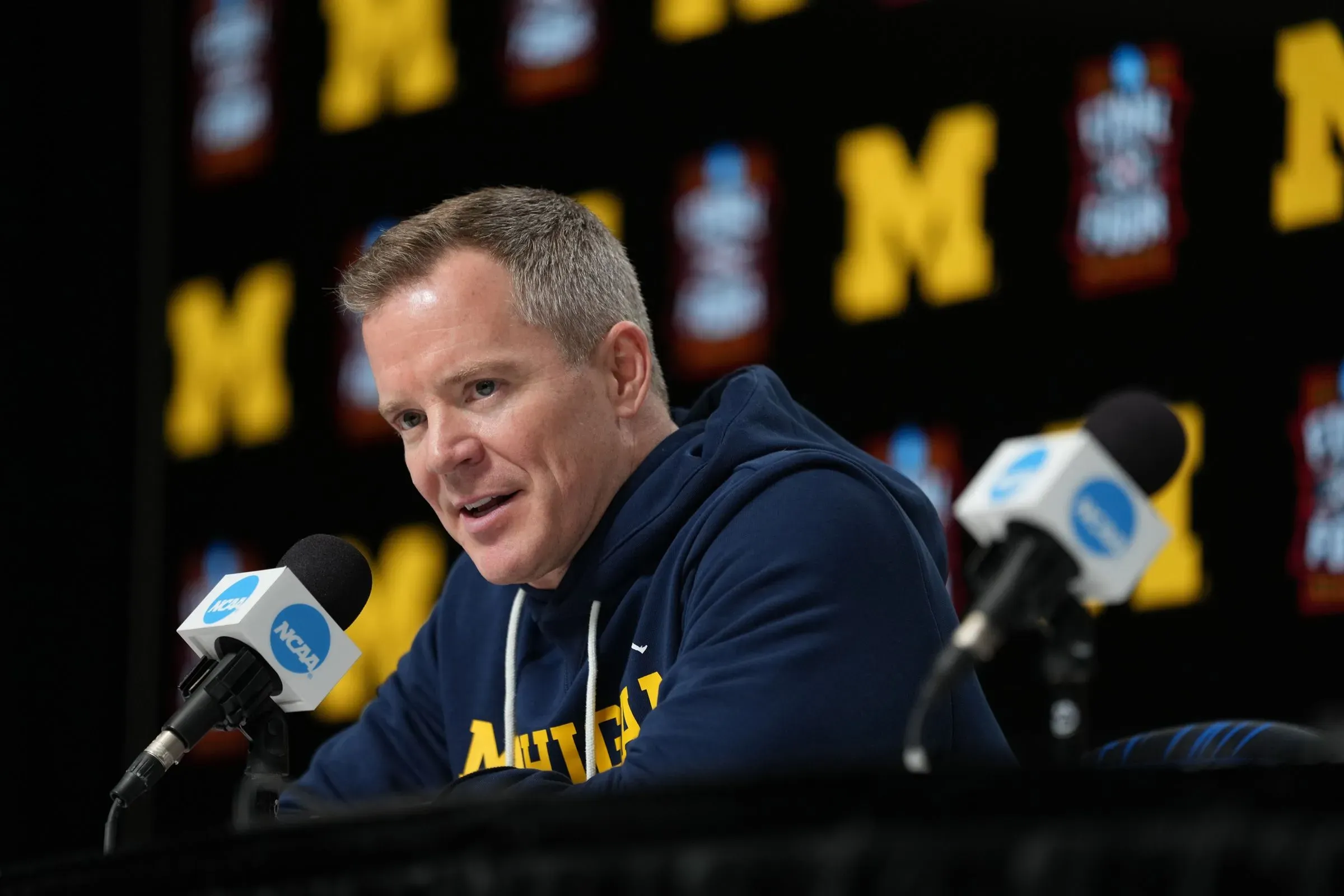 مدرب فريق ولفيرينز، داستي ماي، يتحدث في مؤتمر صحفي، مع خلفية تحمل شعار جامعة ميشيغان، مع التركيز على نجاح الفريق في كرة السلة الجامعية.