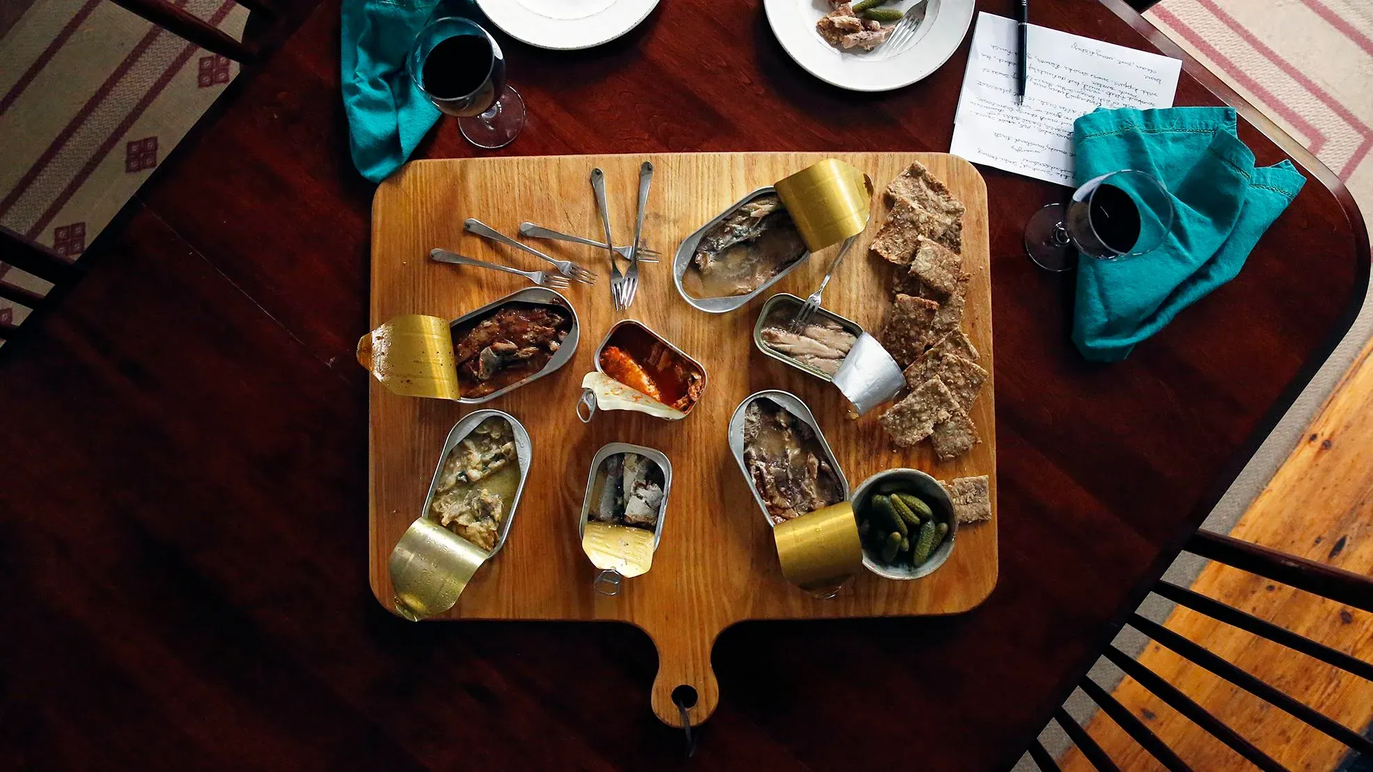 تشكيلة من علب السمك المعلب، تشمل السردين والماكريل، مرتبة بشكل جذاب على لوح خشبي، مع أكواب شراب وأطباق جانبية.