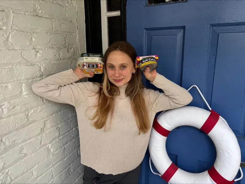 امرأة مبتسمة تحمل علبتين من السمك المعلب، وتقف أمام باب مزين بحلقة نجاة، مما يعكس شعبية الأسماك المعلبة في الثقافة الأمريكية.