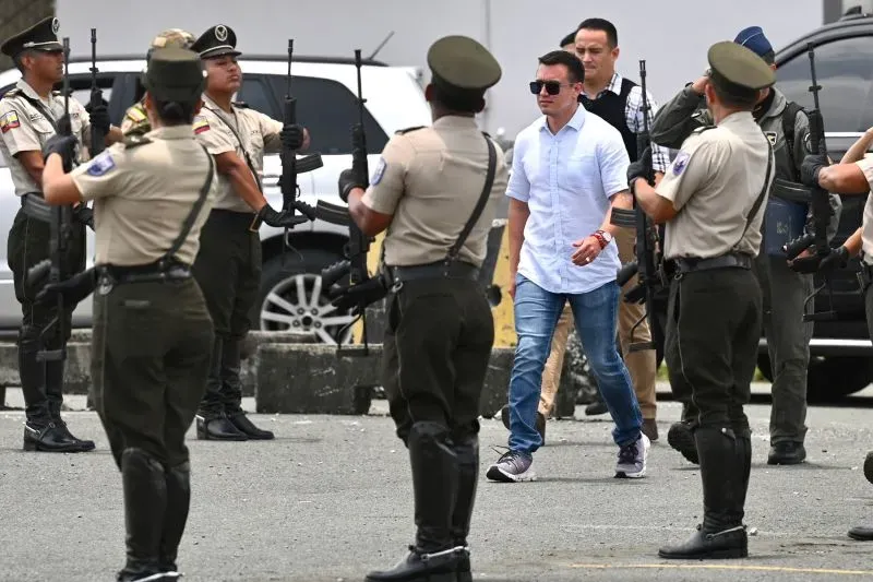 رجل يرتدي قميصًا أبيض يسير بين جنود مسلحين، في سياق أمني متوتر بالإكوادور، وسط تحضيرات لمواجهة العصابات.