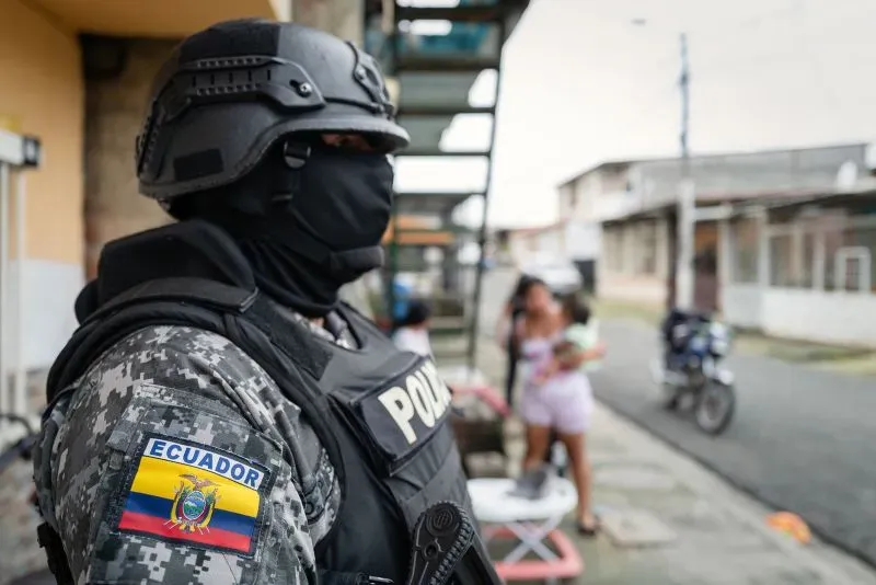شرطي إكوادوري يرتدي زيًا عسكريًا كاملًا، يقف في منطقة حضرية، بينما تظهر نساء في الخلفية، مما يعكس الأوضاع الأمنية المتوترة في البلاد.