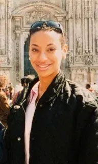 امرأة مبتسمة ترتدي سترة داكنة ونظارات شمسية، تقف أمام معلم تاريخي، تعكس القضايا الإنسانية المتعلقة بالاحتجاز في الصين.