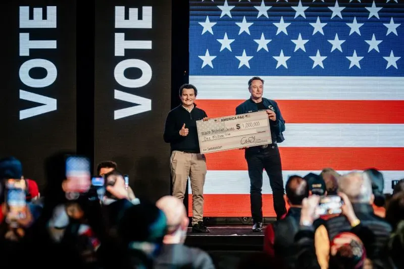 إيلون ماسك يقدم شيكًا بقيمة مليون دولار لأحد الحضور خلال حدث انتخابي في ويسكونسن، مع العلم الأمريكي خلفه، في إطار دعم المرشح المحافظ.