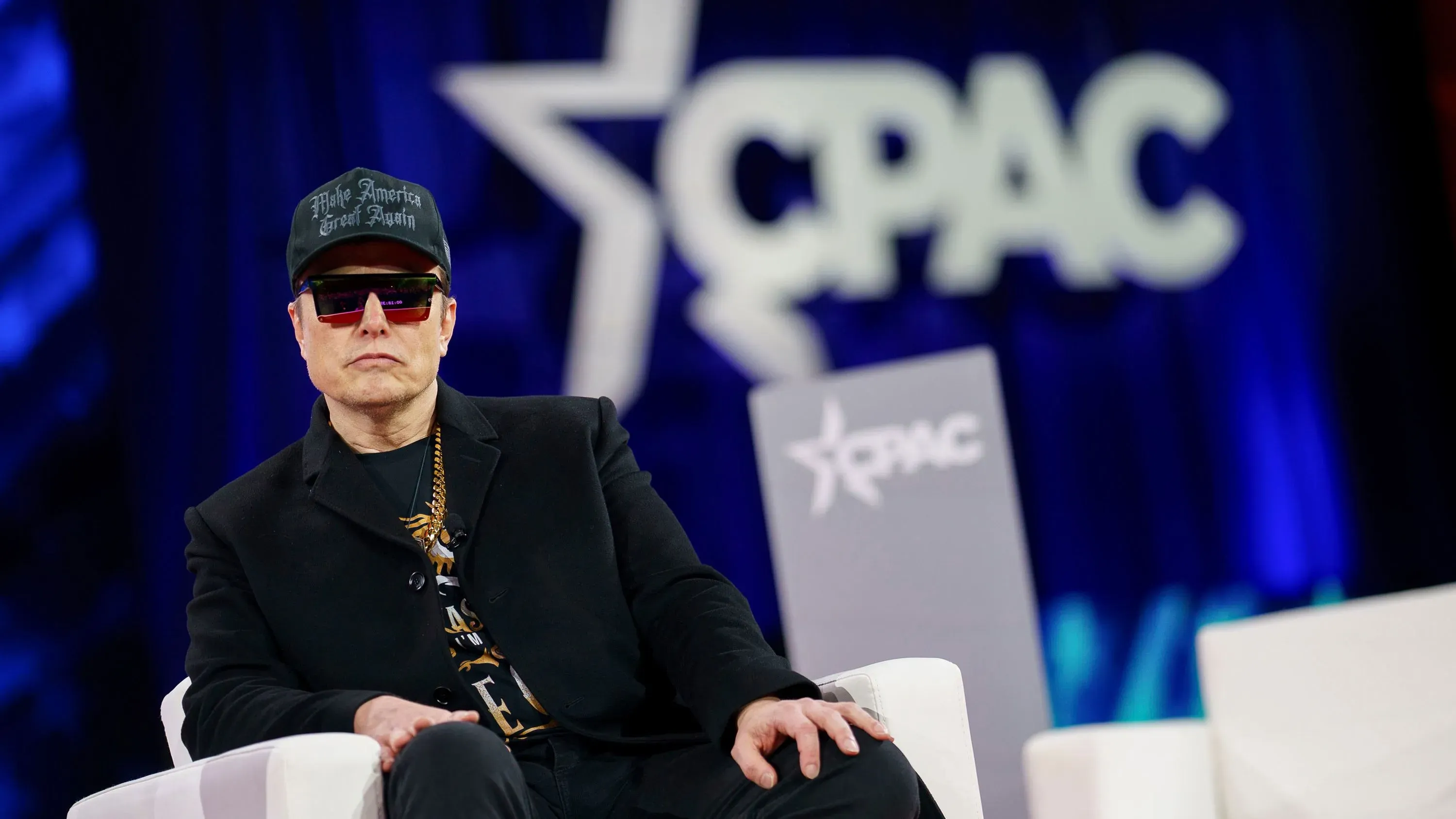 إيلون ماسك يرتدي نظارات شمسية وقبعة خلال حدث، مع وجود شعار CPAC في الخلفية، وسط تراجع أسهم تسلا ومبيعاتها في أوروبا.